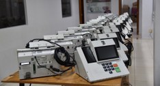 A imagem mostra diversas urnas eletrônicas modelo UE2022 organizadas em duas fileiras sobre mesa...