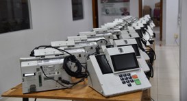 A imagem mostra diversas urnas eletrônicas modelo UE2022 organizadas em duas fileiras sobre mesa...