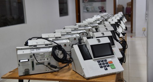 A imagem mostra diversas urnas eletrônicas modelo UE2022 organizadas em duas fileiras sobre mesa...