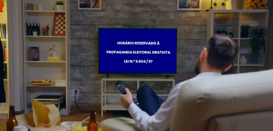Fotografia em que se observa, em primeiro plano e de costas, um homem sentado em um sofá bege, s...